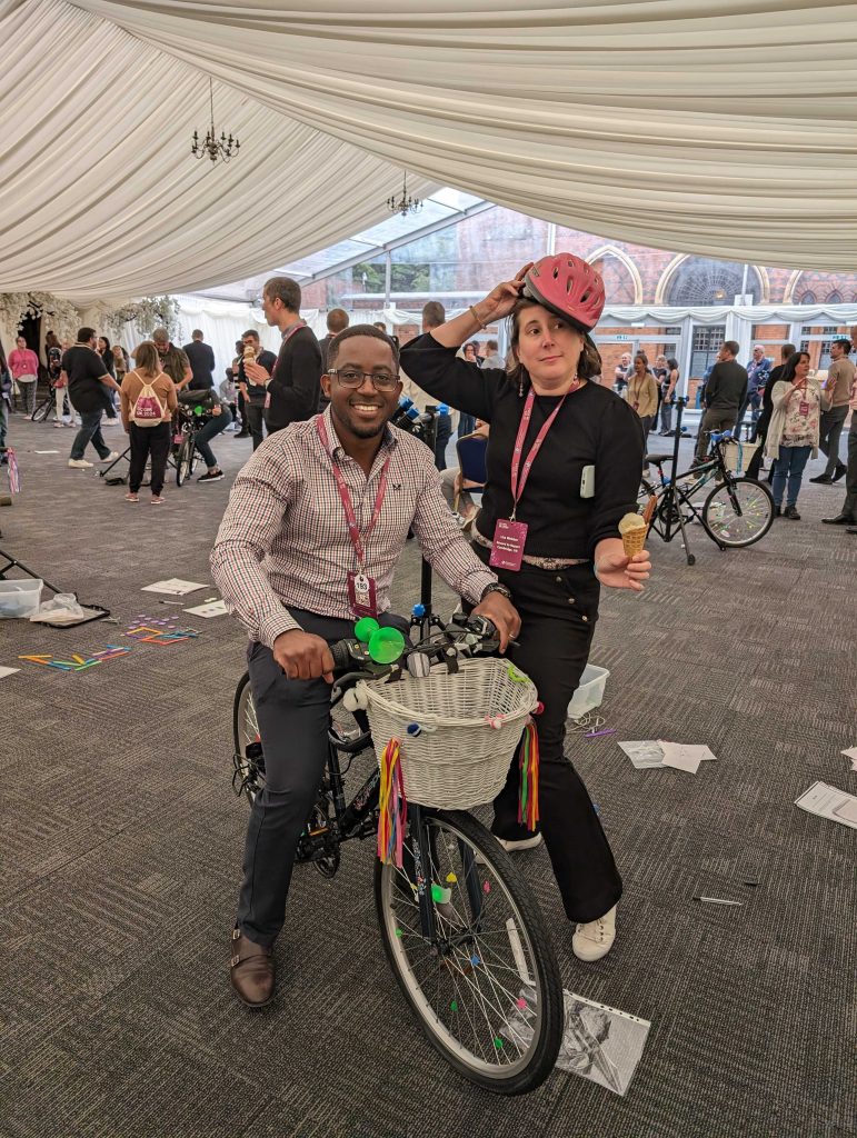 A man smiling and sitting on the bicycle. A woman next to hime posing and smiling with a red hat on