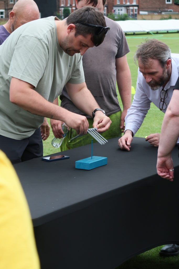 A group of men gathered around a black table outdoors, focused on a balancing challenge involving several forks balanced on a toothpick-like rod anchored in a blue base. One man is adjusting the forks while holding a water bottle, as others observe closely. The activity appears to be part of a team-building or problem-solving exercise on a grassy field.