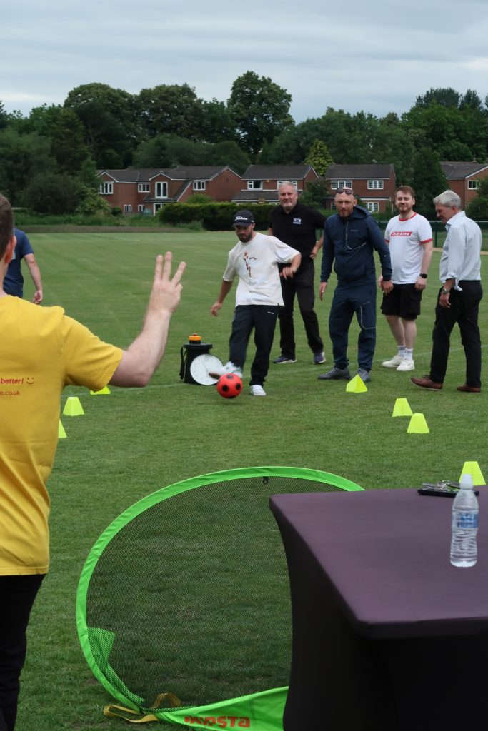 A group of men participating in an outdoor team-building activity on a grassy field. One man in a white shirt and cap is kicking a red football toward a small green goal, while others stand behind him watching and smiling. Yellow cones mark the course, and a person in a yellow shirt with a stopwatch stands in the foreground, signaling with two fingers. Residential houses and trees are visible in the background under a cloudy sky.